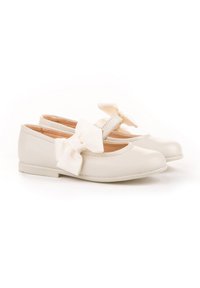 Zapatos tipo Mary Jane para niños en color blanco, con suelas planas y grandes lazos de tela en las correas, presentados sobre un fondo blanco.