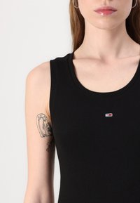 Crni rebrasti majica bez rukava s okruglim izrezom i malim izvezenim logom na prsima. Glatka tekstura i pripijeni dizajn.