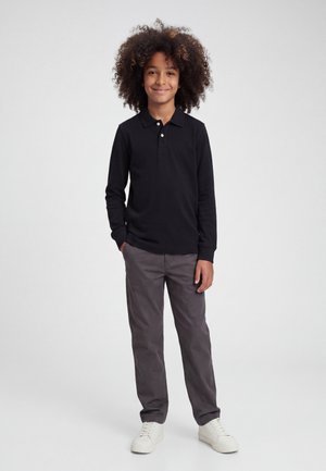 Enfant aux cheveux bouclés portant un polo noir à manches longues, un pantalon gris et des baskets blanches, debout sur un fond clair uni.