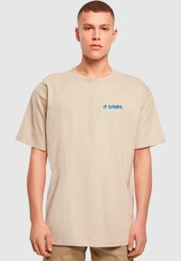 Beige oversized t-shirt i bomuld med et blåt trykt logo på venstre bryst. Korte ærmer med rund halsudskæring og en glat tekstur.