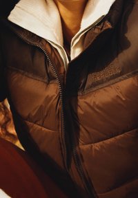 Brun vadderad jacka med vitt fleecefoder och dragkedja. "PACIFIC REPUBLIC"-logotypen är sydd på bröstet. Har synliga sömmar som detalj.