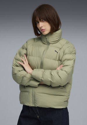 Vrouw met kort bruin haar, gekleed in een lichtgroene pufferjack met een hoge kraag en zwarte broek, met de armen gekruist voor een grijze achtergrond.