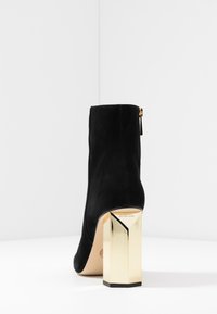 Botín de tobillo de ante negro con un tacón bloque geométrico dorado y un detalle de cremallera lateral. Textura suave. Nombre de la marca en la suela.