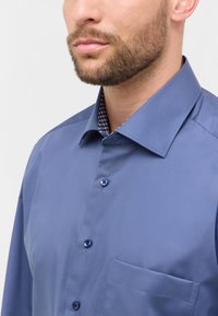 Camicia azzurra chiara con colletto strutturato, taschino sul petto e bottoni blu in contrasto. Il colletto interno presenta una stoffa a fantasia.