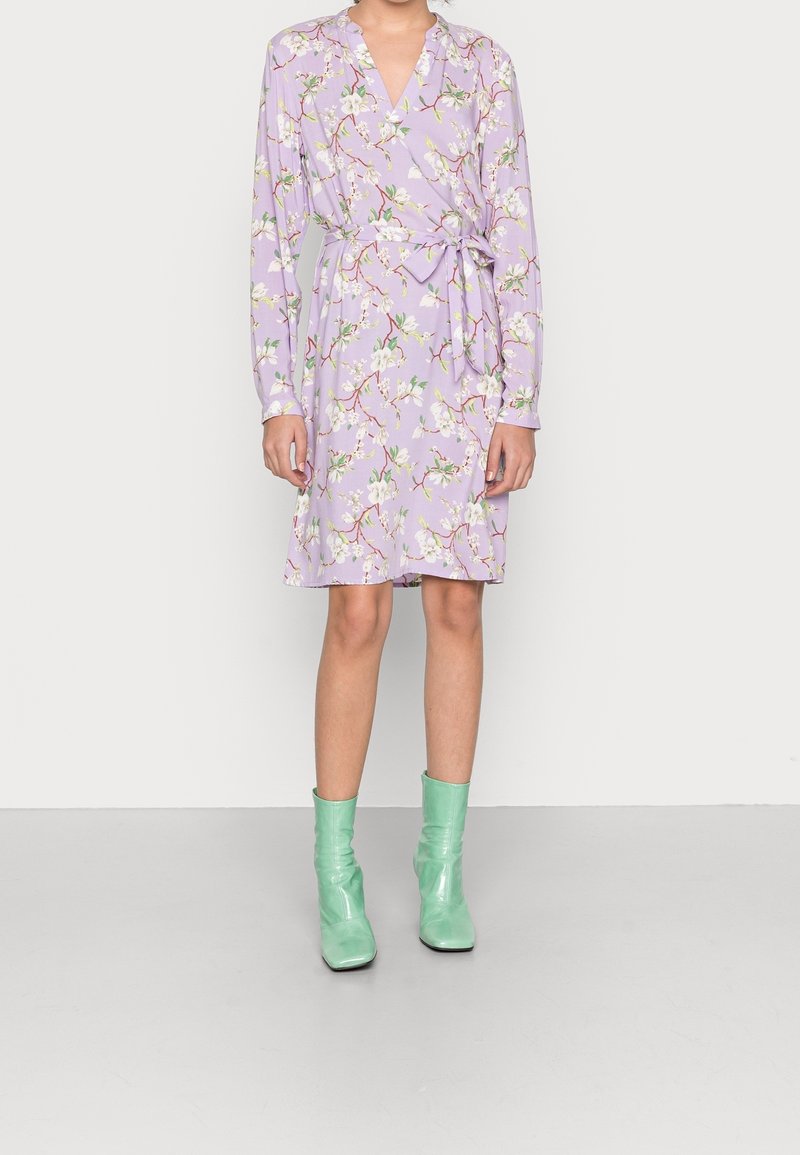 Femme portant une robe portefeuille fleurie lavande à manches longues avec un nœud latéral, associée à des bottines vert menthe, debout sur un fond uni.