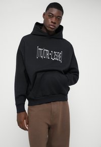 Νεαρός άντρας φοράει μαύρη κουκούλα με λευκό κείμενο "[TTC/CTRL-Z_25.EXE]" και καφέ παντελόνι, που στέκεται μπροστά σε απλό ανοιχτό φόντο.