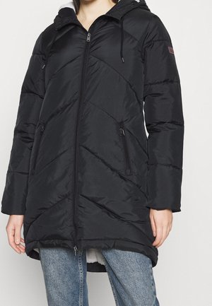 Femme portant une doudoune matelassée noire avec capuche, fermeture éclair à l'avant et poches zippées, associée à un jean en denim bleu.