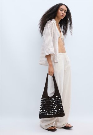 Femme aux longs cheveux bouclés portant une chemise beige rayée, un pantalon blanc ample, des tongs, tenant un sac cabas noir à sequins sur fond clair.