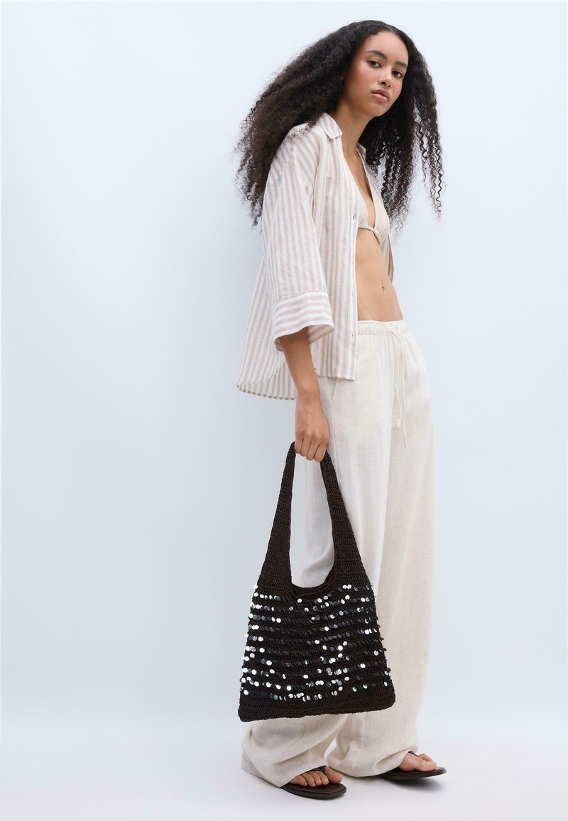 Femme aux longs cheveux bouclés portant une chemise beige rayée, un pantalon blanc ample, des tongs, tenant un sac cabas noir à sequins sur fond clair.