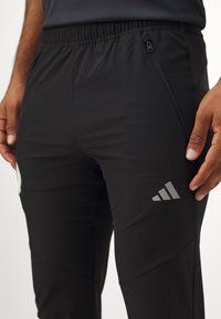 Zwarte sportbroeken met een elastische tailleband, een ritszak en een grijs logo op de linker dij. Glad, lichtgewicht materiaal.