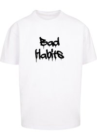 Valkoinen t-paita rentoa istuvuutta, jossa on edessä musta graffiti-tyylinen teksti "Bad Habits", ja se näyttää valuvalta.
