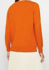 Orangener Pullover mit Rundhalsausschnitt, langen Ärmeln, geripptem Saum und seitlichen Schlitzen. Weiche Textur, getragen über einem plissierten cremefarbenen Rock.