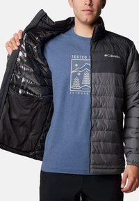 Grau gepolsterte Jacke mit horizontalen Nähten, schwarzem Innenfutter und elastischen Bündchen, getragen über einem blauen T-Shirt mit grafischem Druck.