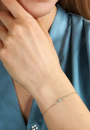Goldfarbene Armbänder mit Kettengliedern, ausgeführt mit zwei horizontalen Stangen, die mit kleinen, funkelnden Kristallen verziert sind. Im Hintergrund ein weicher blauer Seidenstoff.