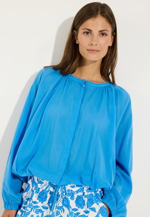 Frau mit glatten braunen Haaren trägt eine lockere hellblaue Bluse und weiße Hosen mit blauem Blumenmuster, steht vor einem weißen Hintergrund.