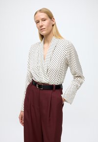 Bluzica s točkicama u krem i crnoj boji, na preklop s dugim rukavima. Uparena s vinscrvenim hlačama s visokim strukom i crnim remenom.