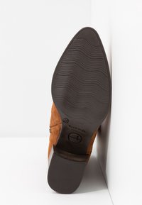 Botte en daim brun à bout pointu avec semelle en caoutchouc noire et petit talon bloc, taille 37, positionnée debout contre un mur blanc.