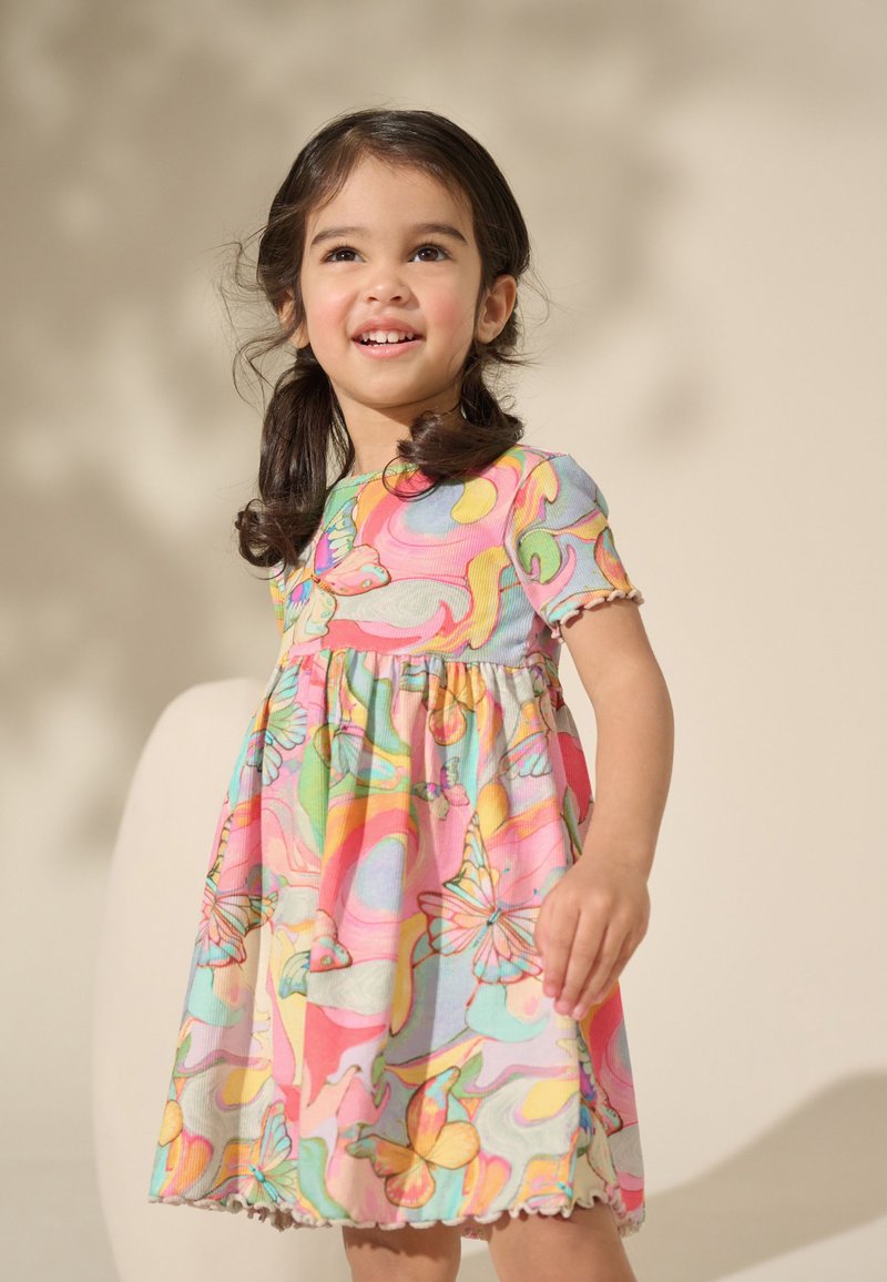 Jeune fille souriante aux cheveux foncés en couettes portant une robe colorée à motif papillons, debout devant un fond beige.