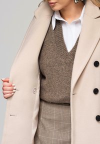 Mujer vestida con abrigo beige, suéter marrón de cuello en V sobre camisa blanca y falda de cuadros, mostrando el forro interior del abrigo con una mano.