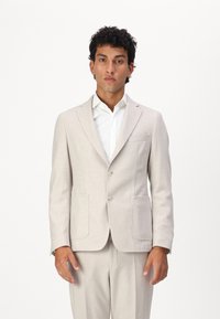 Blazer bej deschis cu două butoane, rever cu tăietură și două buzunare frontale. Purtați peste o cămașă albă, dintr-un material texturat.