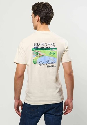 Hombre de espaldas que lleva una camiseta beige con un gráfico colorido del Campeonato de Polo Abierto de EE.UU. en Palm Beaches, Florida y vaqueros azules.