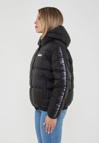 Giacca a vento nera con cappuccio, caratterizzata da un logo lucido sulla manica, texture trapuntata e polsini elasticizzati. Indossata con jeans blu.