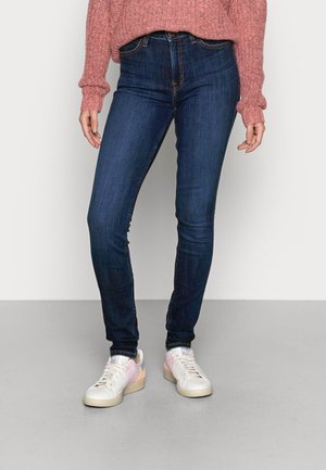 Vrouw draagt donkerblauwe skinny jeans, witte sneakers met roze accenten en een gestructureerde roze trui met lange mouwen, staand op een grijze vloer.