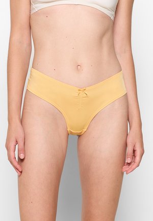 Woman wearing yellow low-rise bikini-style underwear and beige bra, standing against a plain light background.