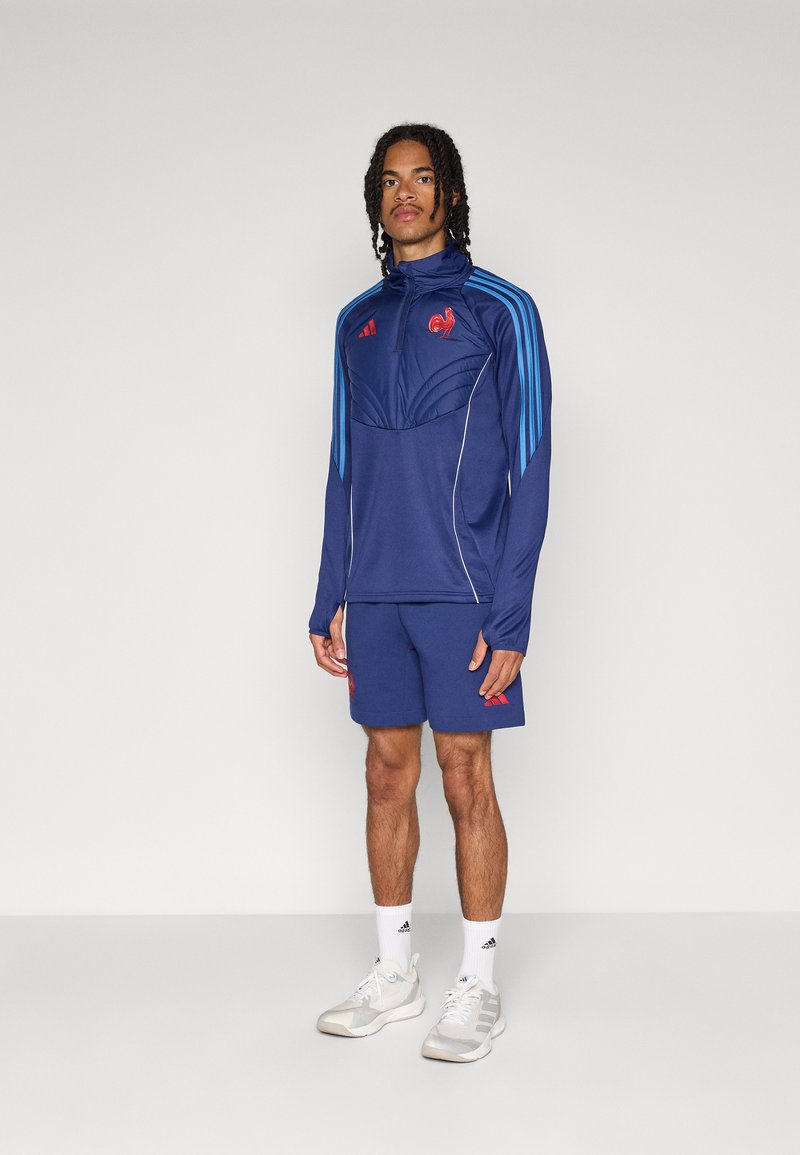 Ensemble de sportswear bleu marine comprenant une veste zippée avec des rayures bleues et des accents rouges, assortie à un short. Fabriqué en tissu respirant.