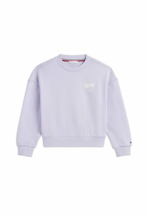 Ung person med krøllet blondt hår iført en lyseblå sweatshirt, mørke bukser og hvide sneakers stående på en hvid baggrund.