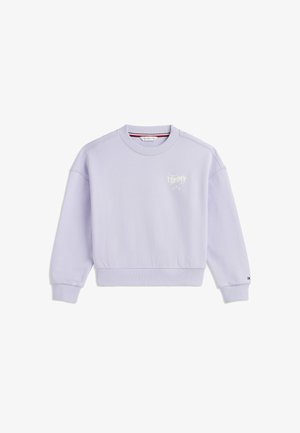 Ung person med krøllet blondt hår iført en lyseblå sweatshirt, mørke bukser og hvide sneakers stående på en hvid baggrund.