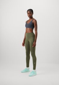 Sötét szürke sportmelltartó, magas derekú olíva zöld leggings, és világos kék sportcipők. Simább textúrák minimalista design kiegészítőkkel.