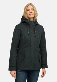 Vaalea nainen, joka on pukeutunut tummanharmaaseen huppariin, jossa on nyörit, etutaskut, napitus sekä sinisiin farkkuihin, seisoo valkoista taustaa vasten.