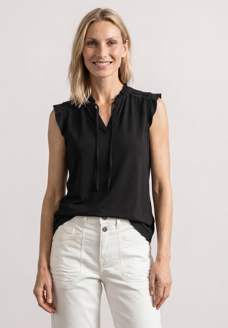 Femme souriante aux cheveux blonds portant un top noir sans manches et un pantalon blanc taille haute, debout devant un fond clair uni.