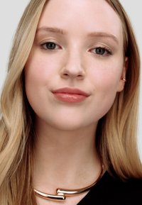 Gulltonet halskjede med en buet design liggende mot en models hals, kombinert med langt, bølgete blondt hår og naturlig sminke.