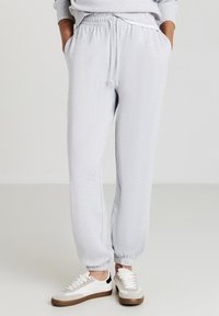 Helle graue Jogginghose mit elastischem Bund und Kordelzug. Mit Bündchen an den Knöcheln und seitlichen Taschen. Kombiniert mit weißen und beigen Turnschuhen.