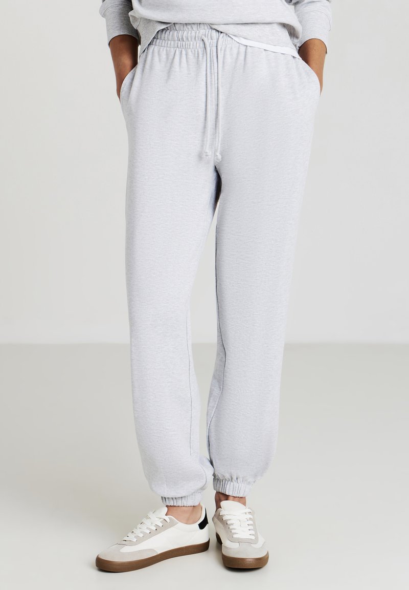 Helle graue Jogginghose mit elastischem Bund und Kordelzug. Mit Bündchen an den Knöcheln und seitlichen Taschen. Kombiniert mit weißen und beigen Turnschuhen.