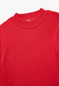 Czerwony sweter z prążkowanej dzianiny o wysokim kołnierzu i teksturowanej tkaninie. Zbliżenie podkreśla jego rozciągliwy, drobno dzianinowy fason.