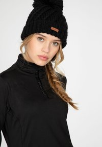 Gorro de lana negro con un pompón en la parte superior y un parche de piel con la marca. Modelo llevando una camiseta de manga larga ajustada negra con un alto cuello.