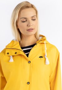 Chaqueta de lluvia impermeable amarilla con capucha, que presenta borlas blancas, un logo de paraguas negro y un zipper plateado. Forro interior a rayas en blanco y negro.