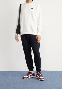 Vit sweatshirt med svart Nike-logga, kombinerad med svarta joggers och vinröda och vita sneakers. Inkluderar en liten svart axelremsväska.