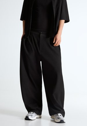 Osoba v voľných čiernych textúrovaných nohaviciach a zodpovedajúcom topu, spárovaná s bielo-sivými teniskami, na jednoduchom pozadí.