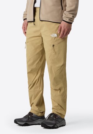 Homme portant un pantalon cargo beige avec des poches zippées et des chaussures de randonnée noires, veste polaire beige visible à la taille.
