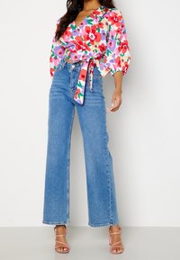 Haut en fleurs avec des manches bouffantes, arborant des motifs rouges, violets et verts ; associé à un jean large taille haute bleu clair et des sandales à talons roses.