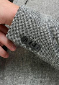 Manica di un cappotto in tessuto grigio testurizzato con quattro bottoni neri. In primo piano si vede una mano che afferra il polsino, evidenziando il dettaglio dei bottoni.