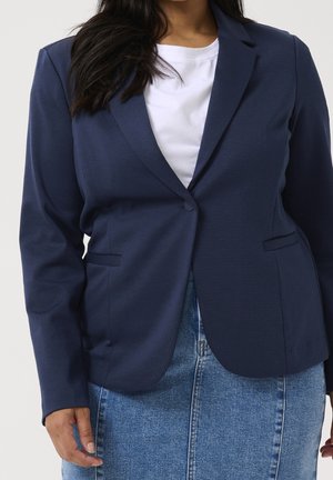 Žena v tmavě modrém saku přes bílou košili a modrou džínovou sukni stojí před jednobarevným pozadím.