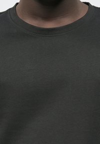 Detailní záběr osoby v jednoduchém tmavě šedém tričku s kulatým výstřihem, které odhaluje krk a horní část hrudníku.