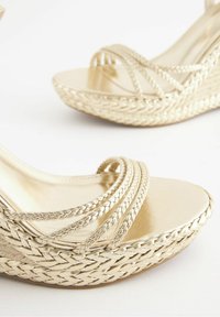 Sandales compensées tressées dorées avec plusieurs fines brides sur les orteils sur fond blanc.