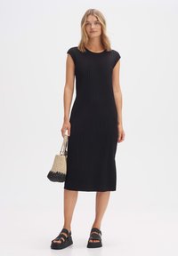 Zwarte geribbelde midi-jurk met kapmouwen, met een getailleerd ontwerp. Gecombineerd met een geweven tas en stevige zwarte sandalen voor contrast.