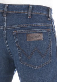 Jeans de mezclilla azul oscuro con una etiqueta de cuero marrón en la pretina con "Wrangler" grabado. Incluye dos bolsillos traseros con costuras en contraste.